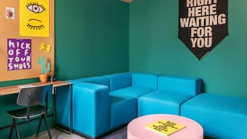 A room with a teal wall, a blue sofa, a pink table, and a bulletin board with various notes and a cactus.