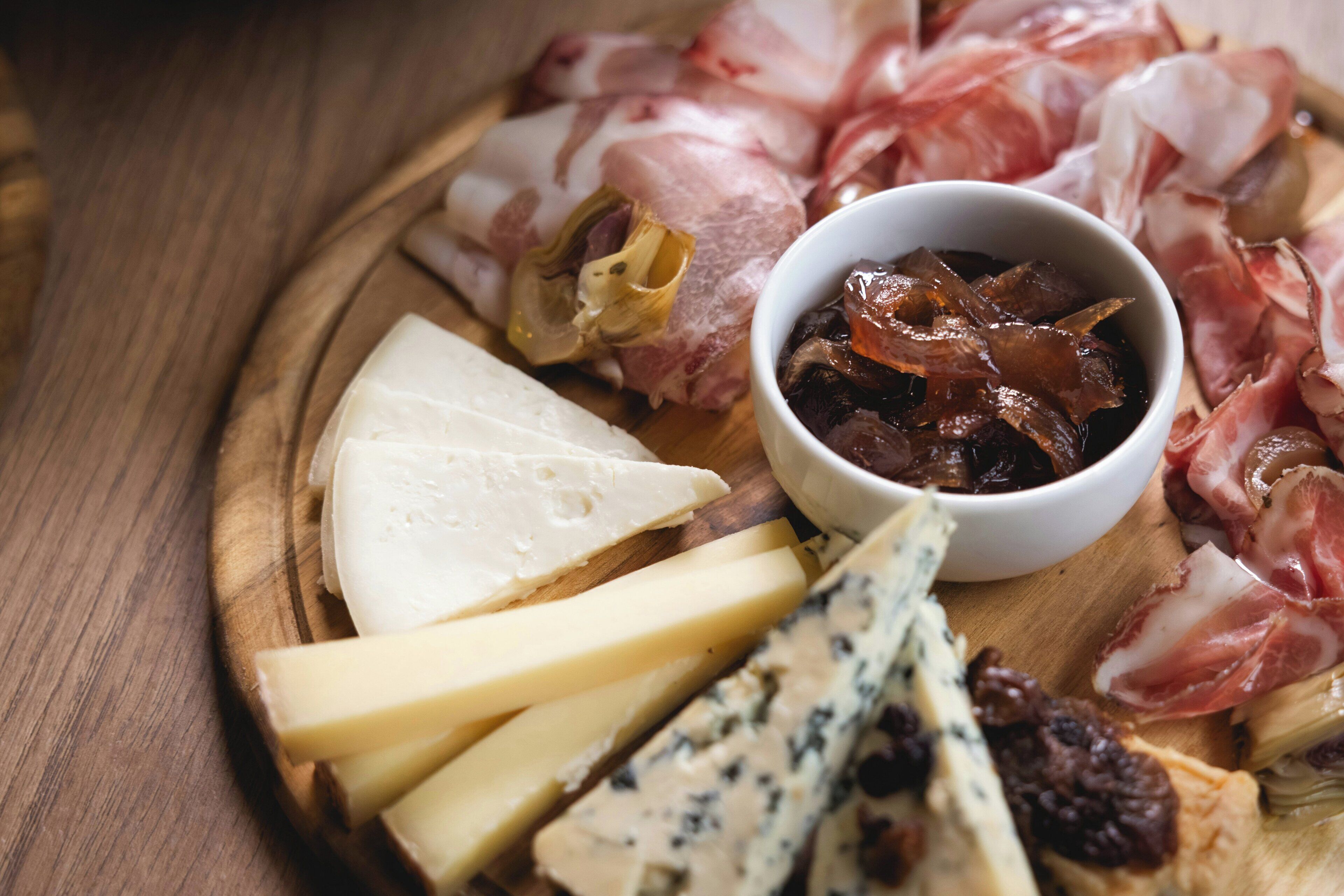 A wooden board with various cheeses, cured meats, and a bowl of pickles.