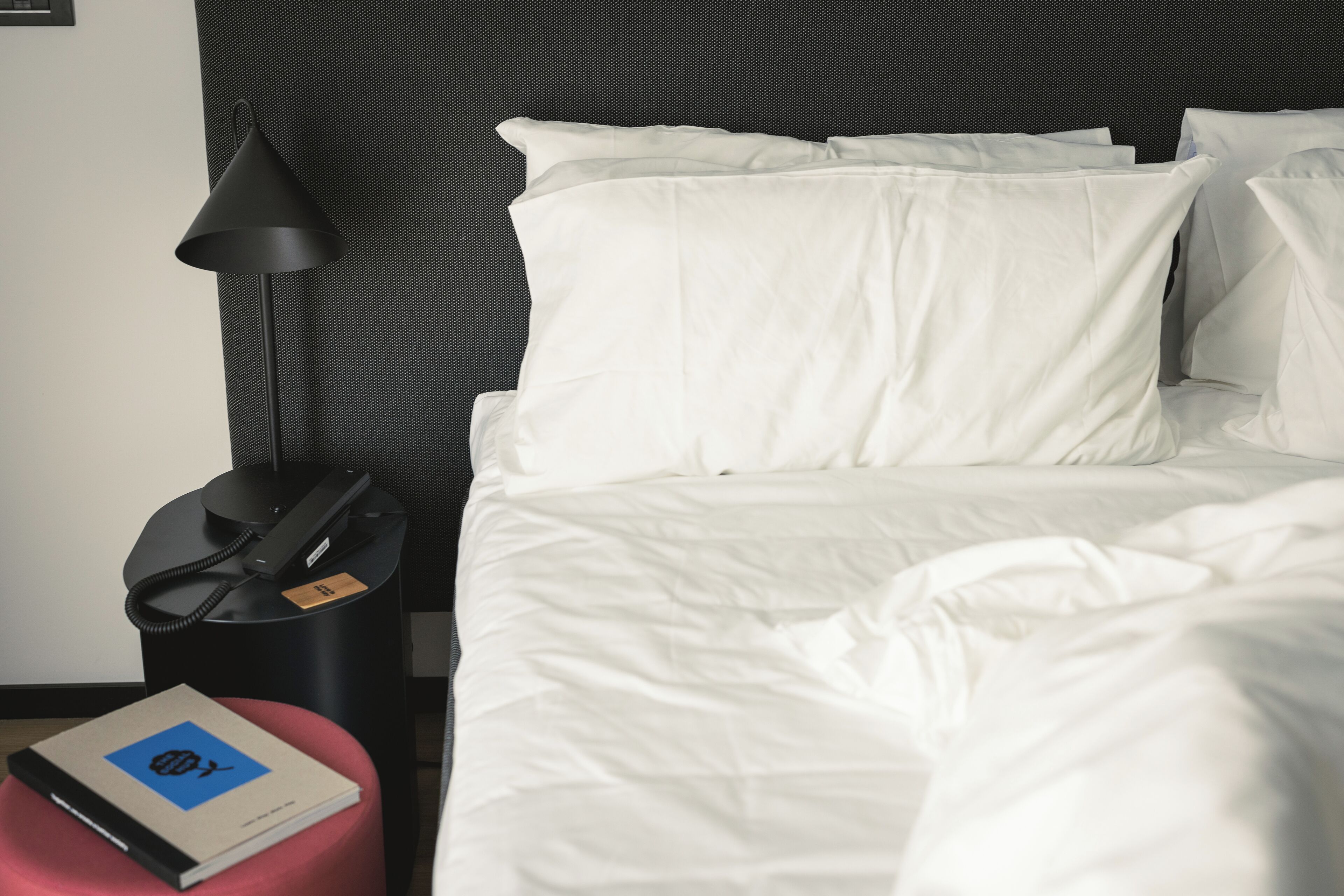 A neatly made bed with white bedding, a black lamp, a telephone, and a book on a red ottoman.