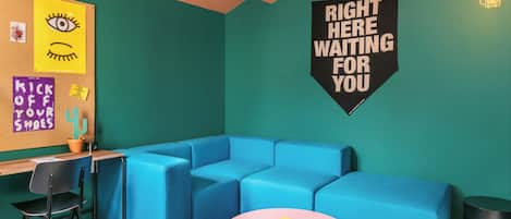 A modern room with a teal wall, a blue sectional sofa, a pink round table, and a bulletin board with various notes and a sign that says 'RIGHT HERE WAITING FOR YOU'.