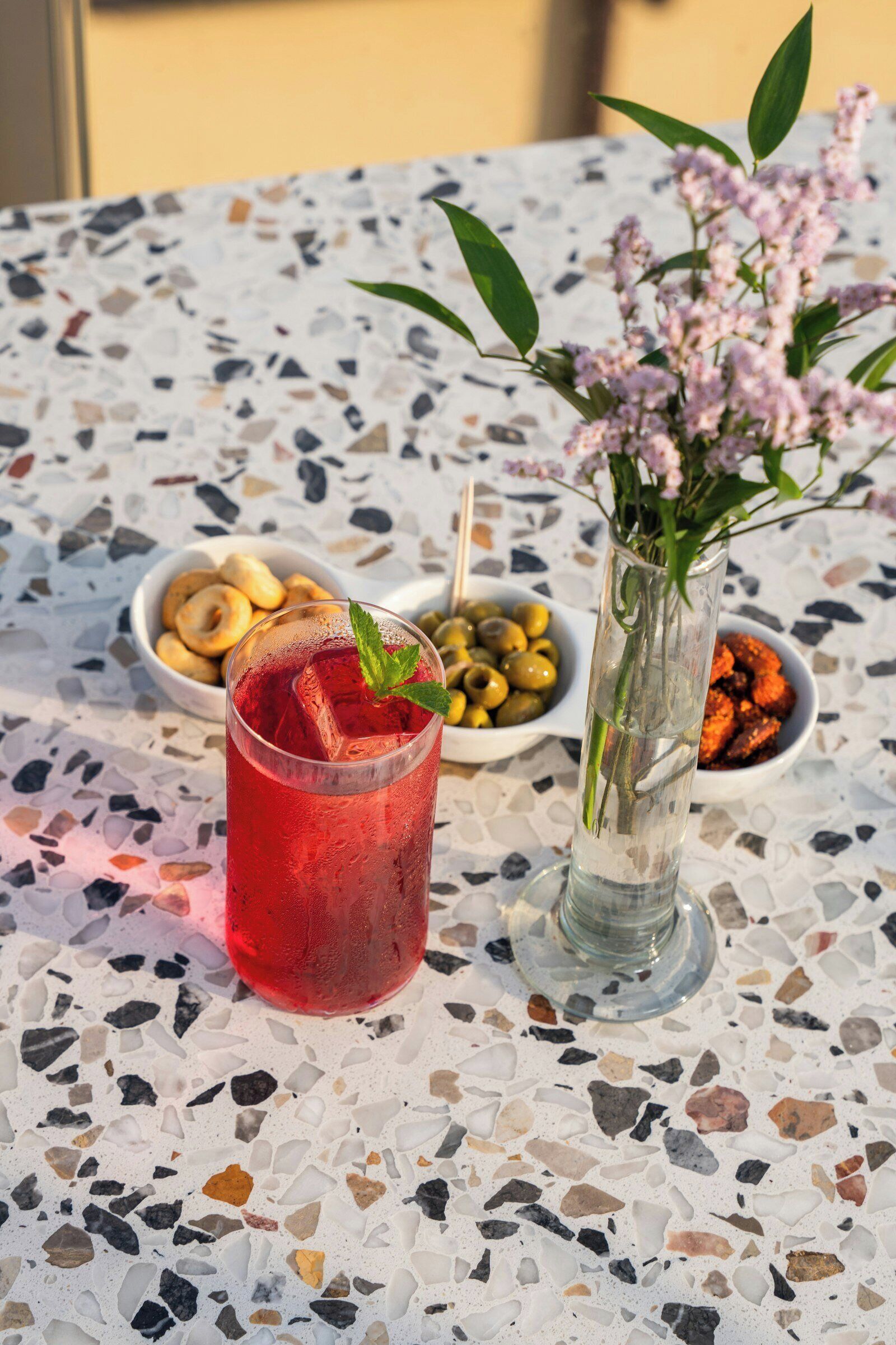 A table with a glass of red beverage, olives, and a vase with flowers.