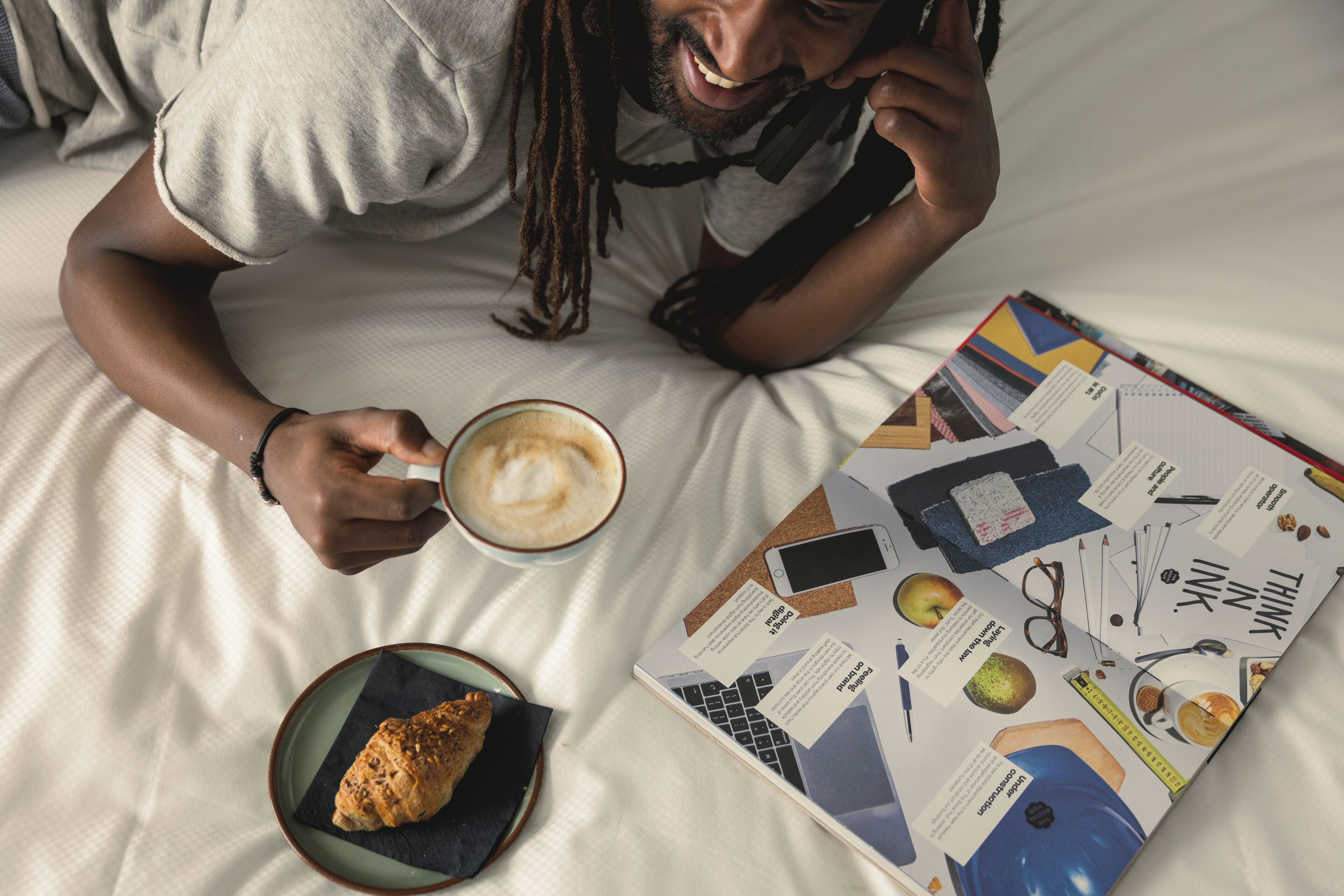 Una persona sdraiata su un letto con un libro, una tazza di caffè e un dolce.
