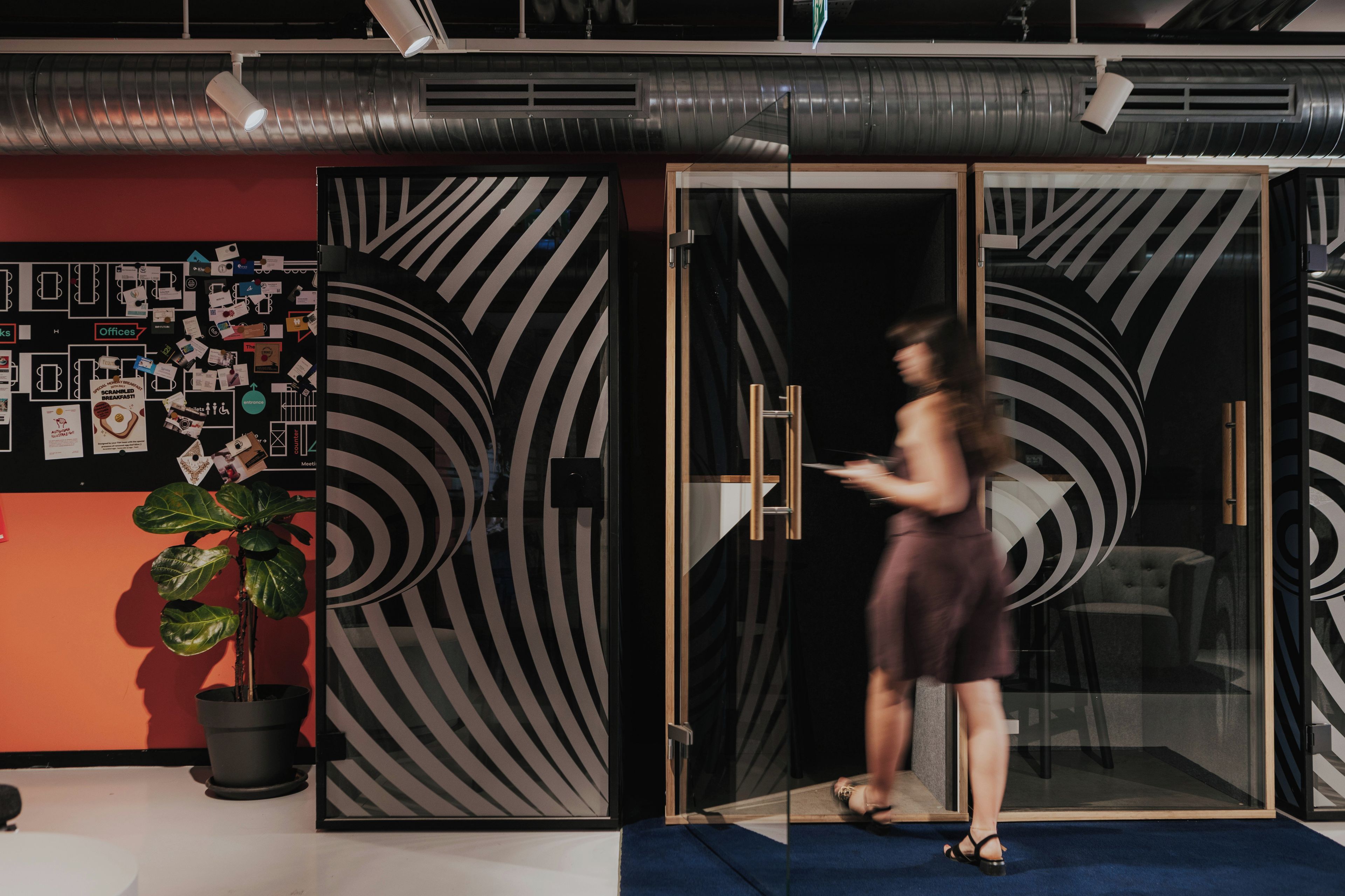 Un espacio de oficina moderno con una puerta de vidrio, un tablón de anuncios con diversos avisos y una planta en maceta.
