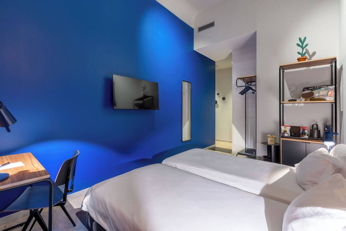 Ein Hotelzimmer mit blauer Akzentwand, einem Bett, einem Schreibtisch mit Stuhl, einem Fernseher und einem Regal mit verschiedenen Gegenständen.