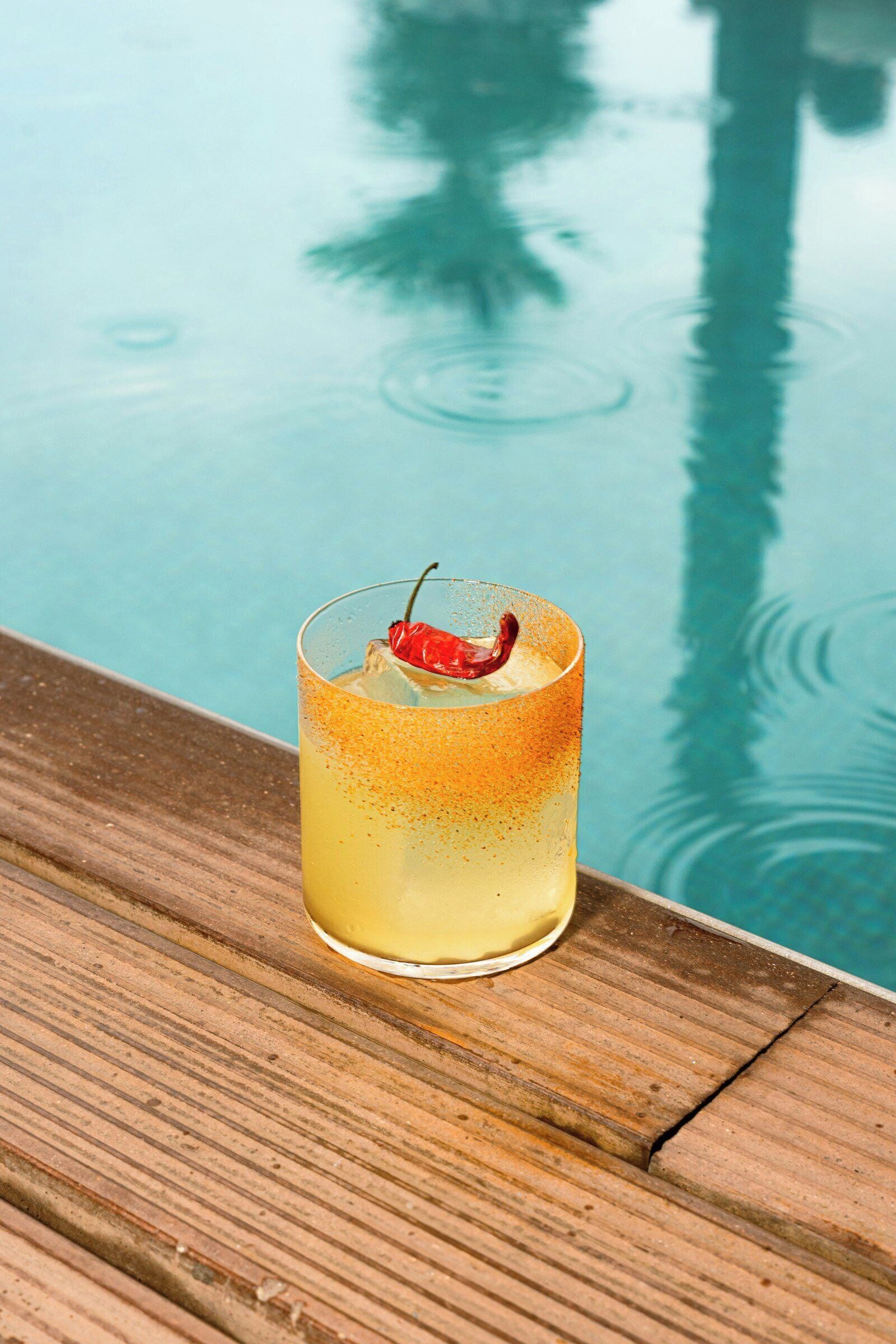 A cocktail with a garnish on a wooden surface near a pool.