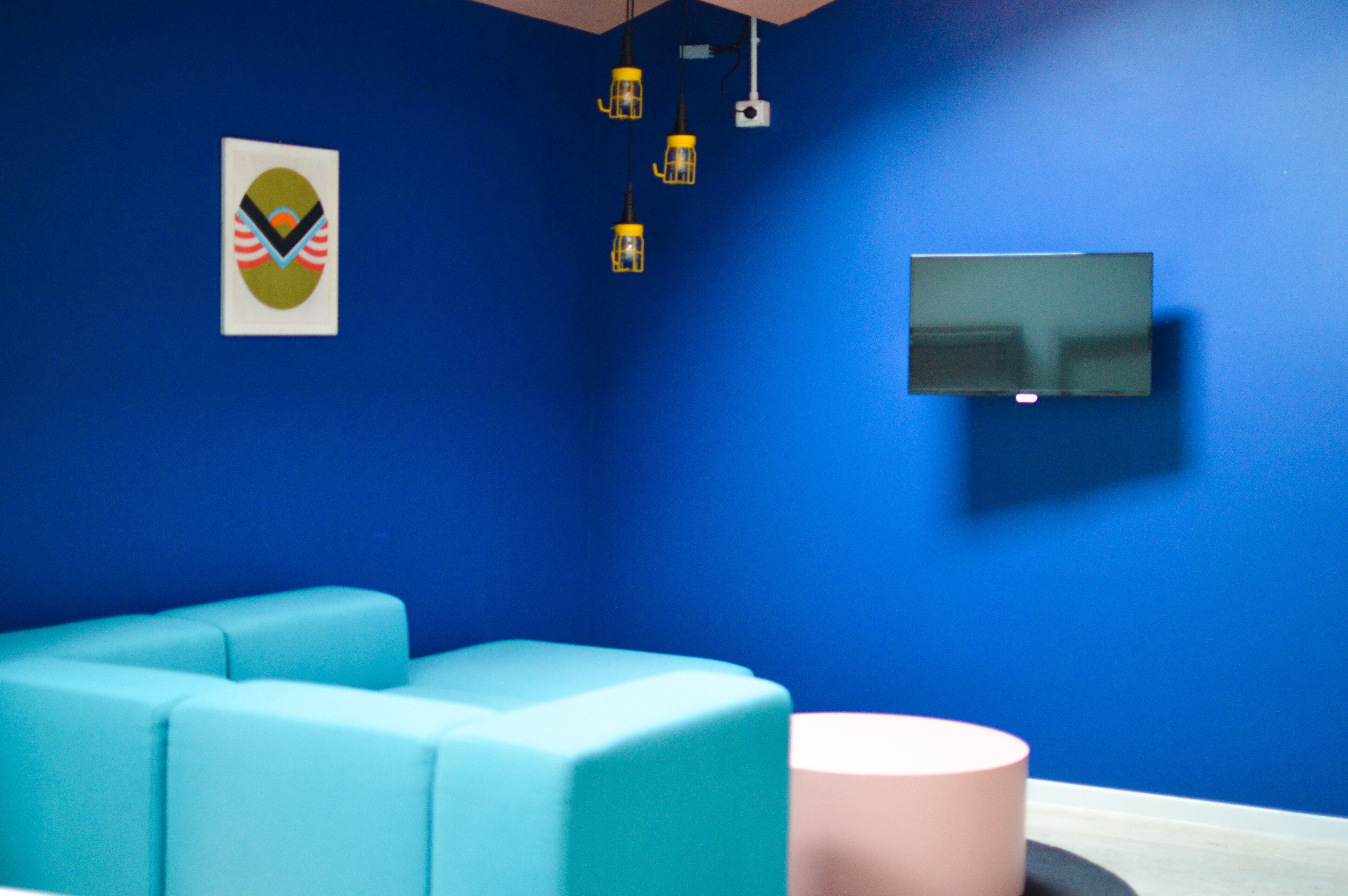 A room with a blue wall, a flat-screen TV mounted on it, a teal sofa, a pink ottoman, and three hanging pendant lights.