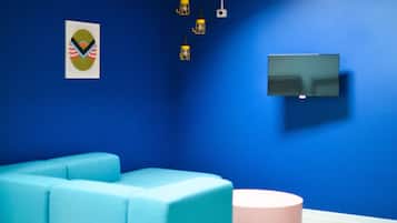 A room with a blue wall, a flat-screen TV mounted on it, a teal sofa, a pink ottoman, and three hanging pendant lights.