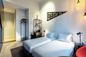 A modern hotel room with a large bed, a wardrobe, a desk, and a 'LET'S STAY IN' sign.