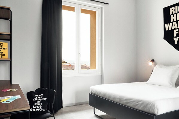 Un dormitorio con cama, escritorio, silla, ventana con cortinas y una lámpara fijada a la pared.