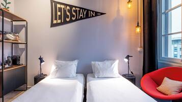 Habitación de hotel con dos camas, una silla roja, una estantería y un cartel que dice 'LET'S STAY IN'.