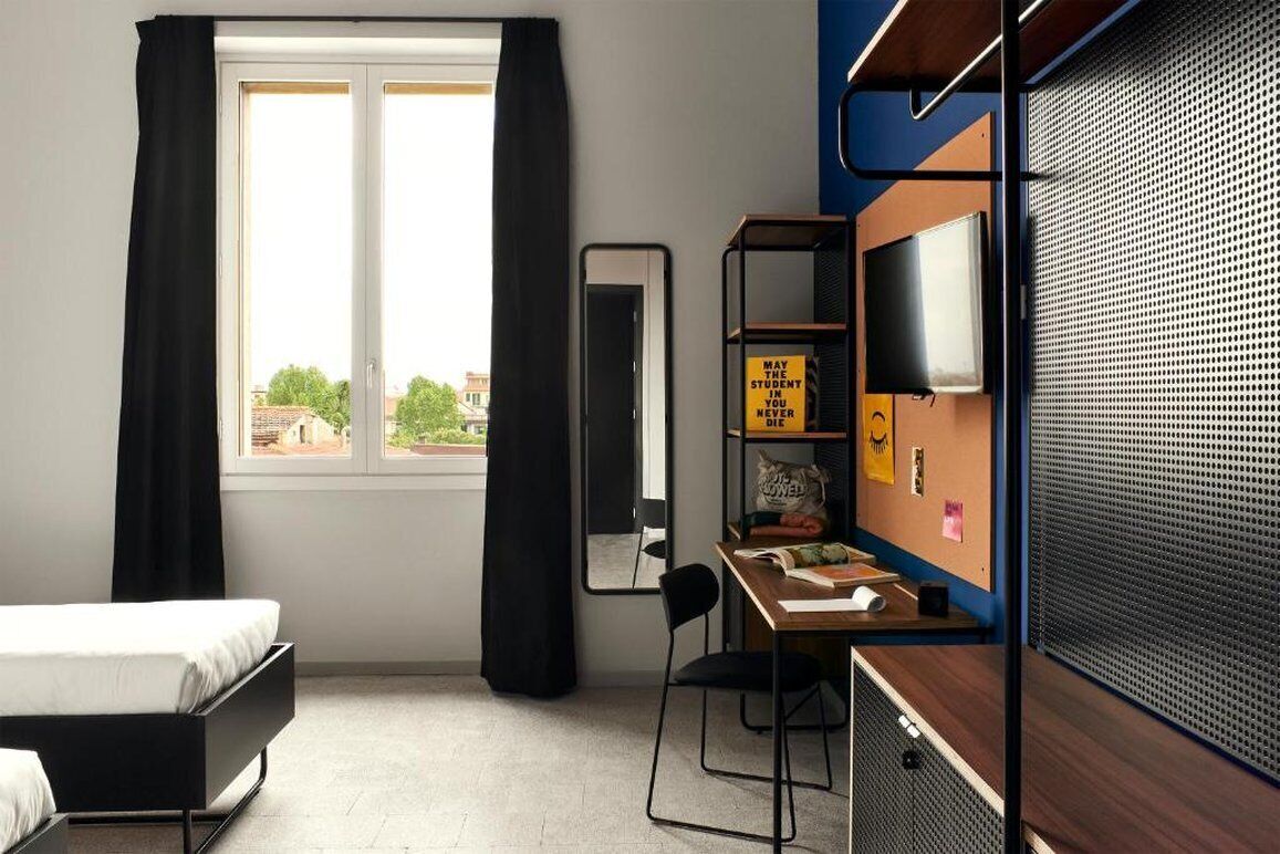 Habitación moderna con cama, escritorio, silla, espejo, televisión y ventana con cortinas.