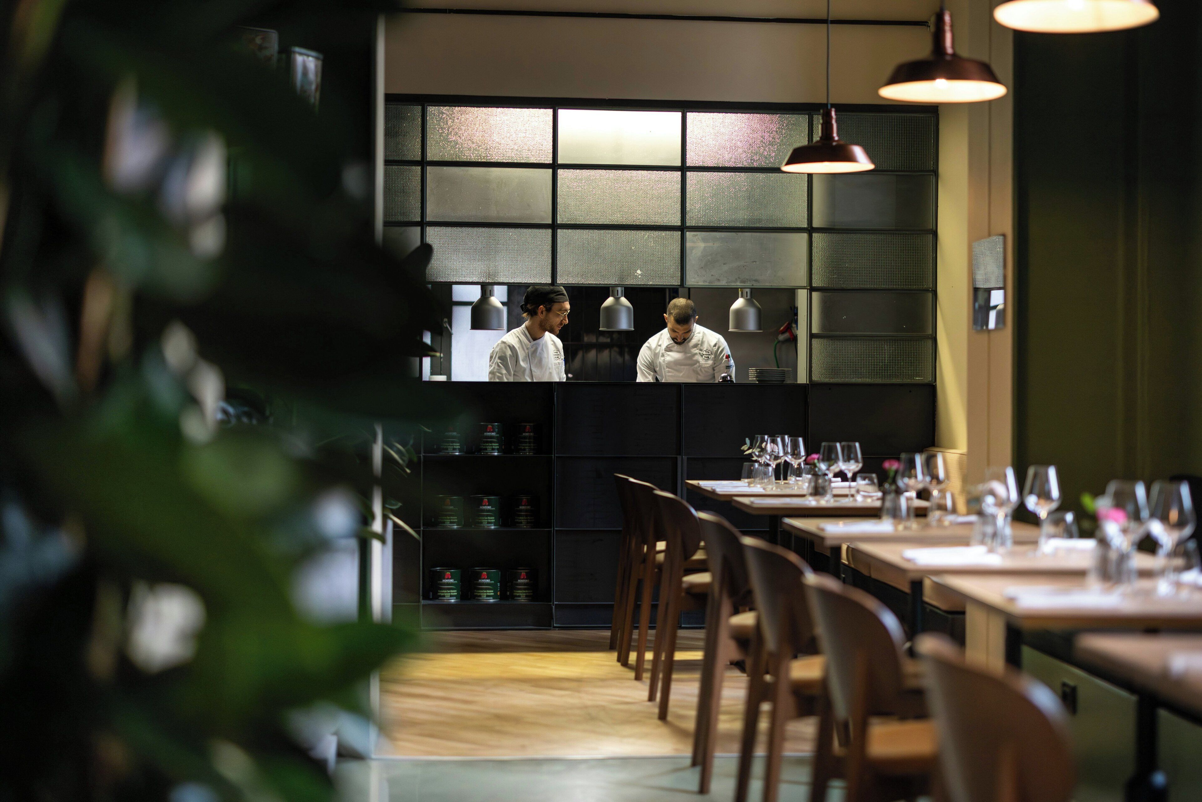 A restaurant interior with a chef at the counter, wooden tables, and chairs.