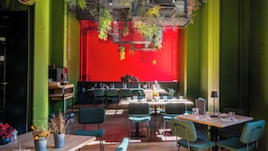 Un restaurante moderno con paredes verdes, detalles en rojo y plantas colgantes. Las mesas están dispuestas con sillas, y hay plantas sobre ellas.
