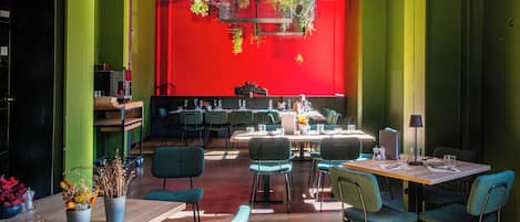 Un restaurant moderne aux murs verts, agrémenté de touches de rouge et de plantes suspendues. Les tables sont disposées avec des chaises, et des plantes sont également placées sur les tables.