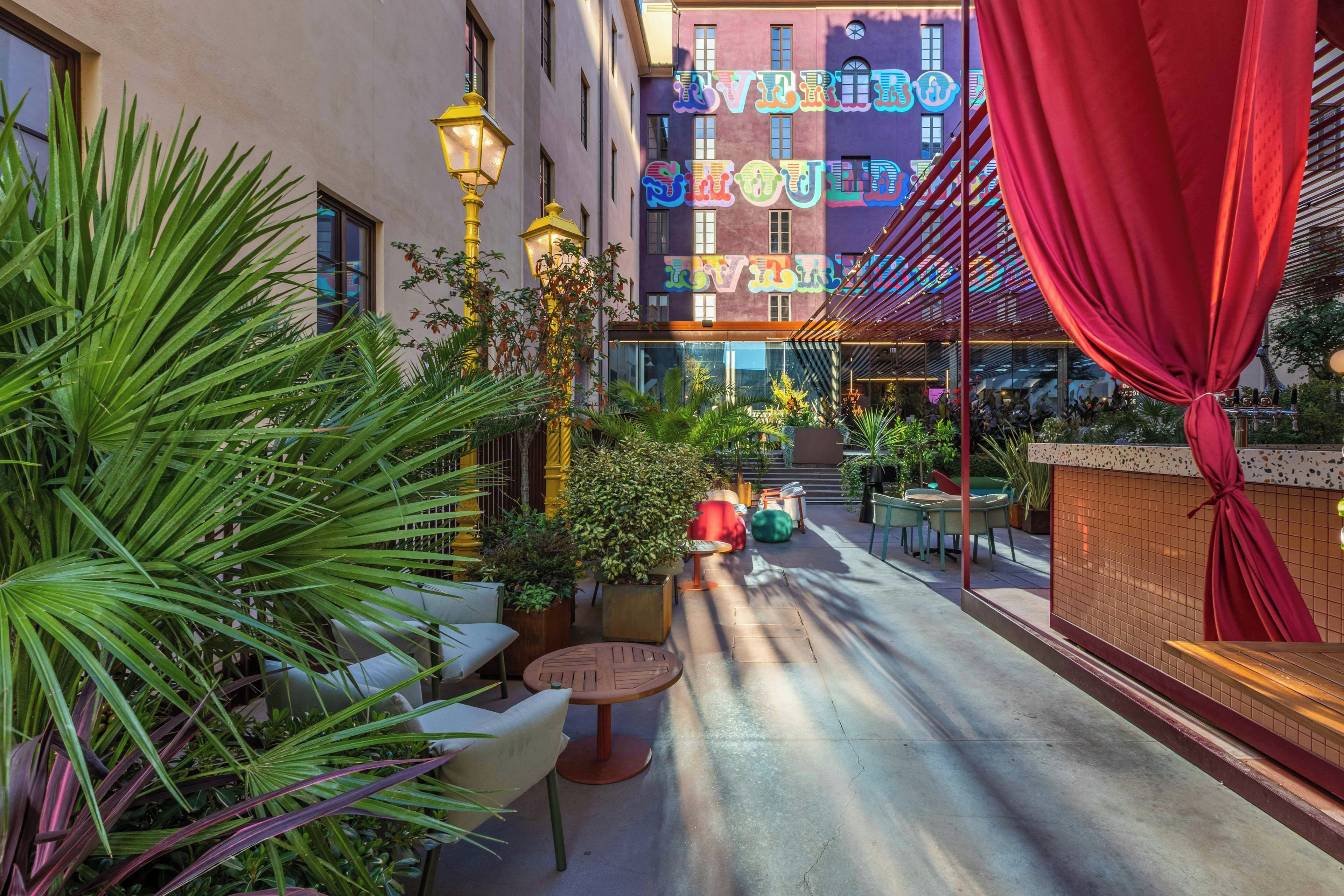 An outdoor seating area with tables and chairs, surrounded by plants and a building with colorful signage.