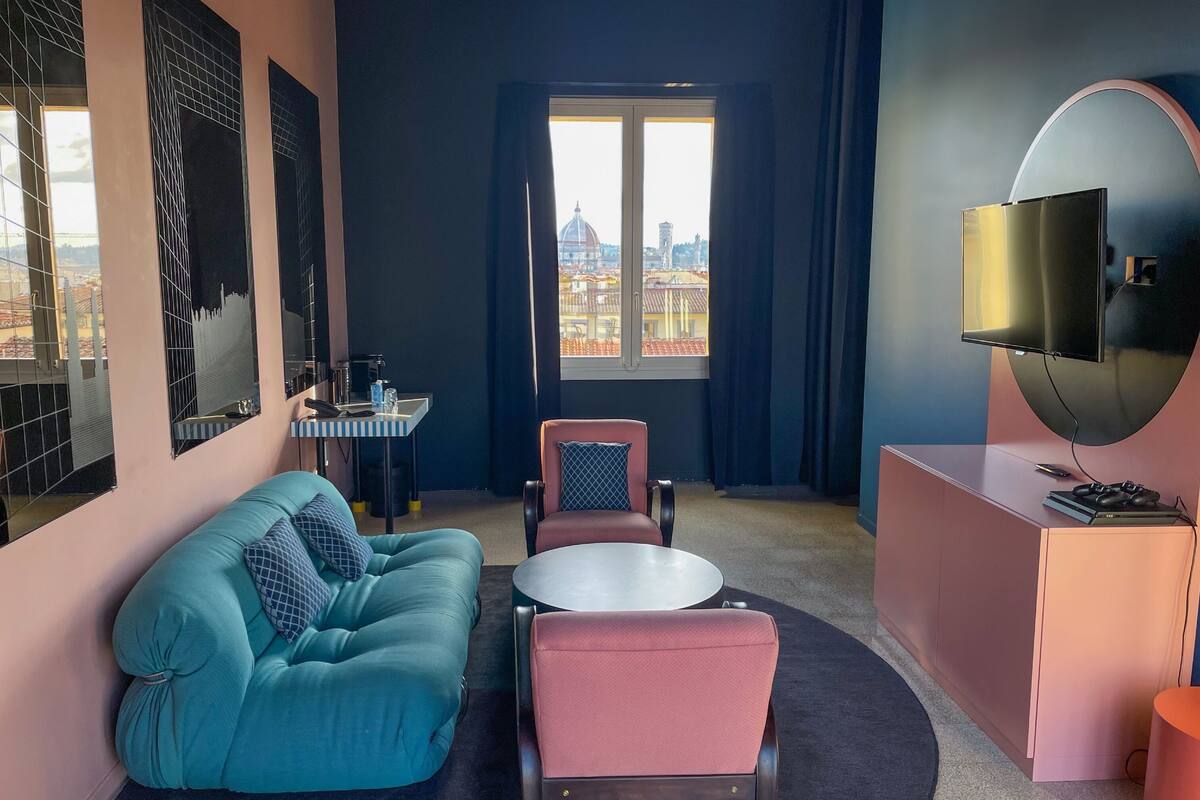 Ein modernes Hotelzimmer mit einem türkisfarbenen Sofa, einem roten Sessel, einem kleinen runden Tisch, einem an einer gewölbten Wand montierten Fernseher und einem Fenster mit Blick auf die Stadt.