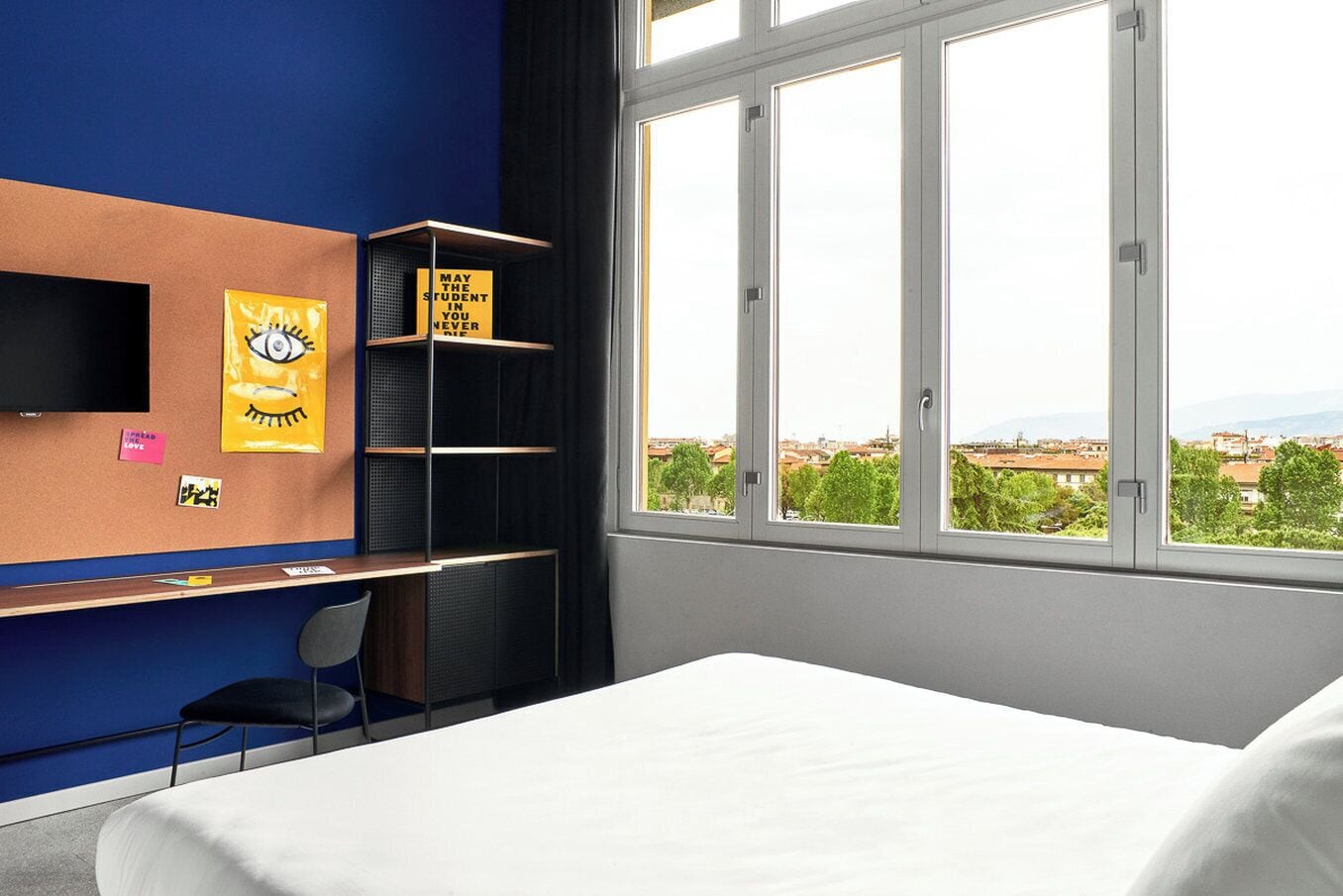 Une chambre d’hôtel comprenant un lit, un bureau avec une chaise, un téléviseur fixé au mur et une fenêtre donnant sur la ville.