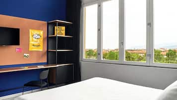 Quarto de hotel com cama, escrivaninha com cadeira, televisão na parede e janela com vista para a cidade.