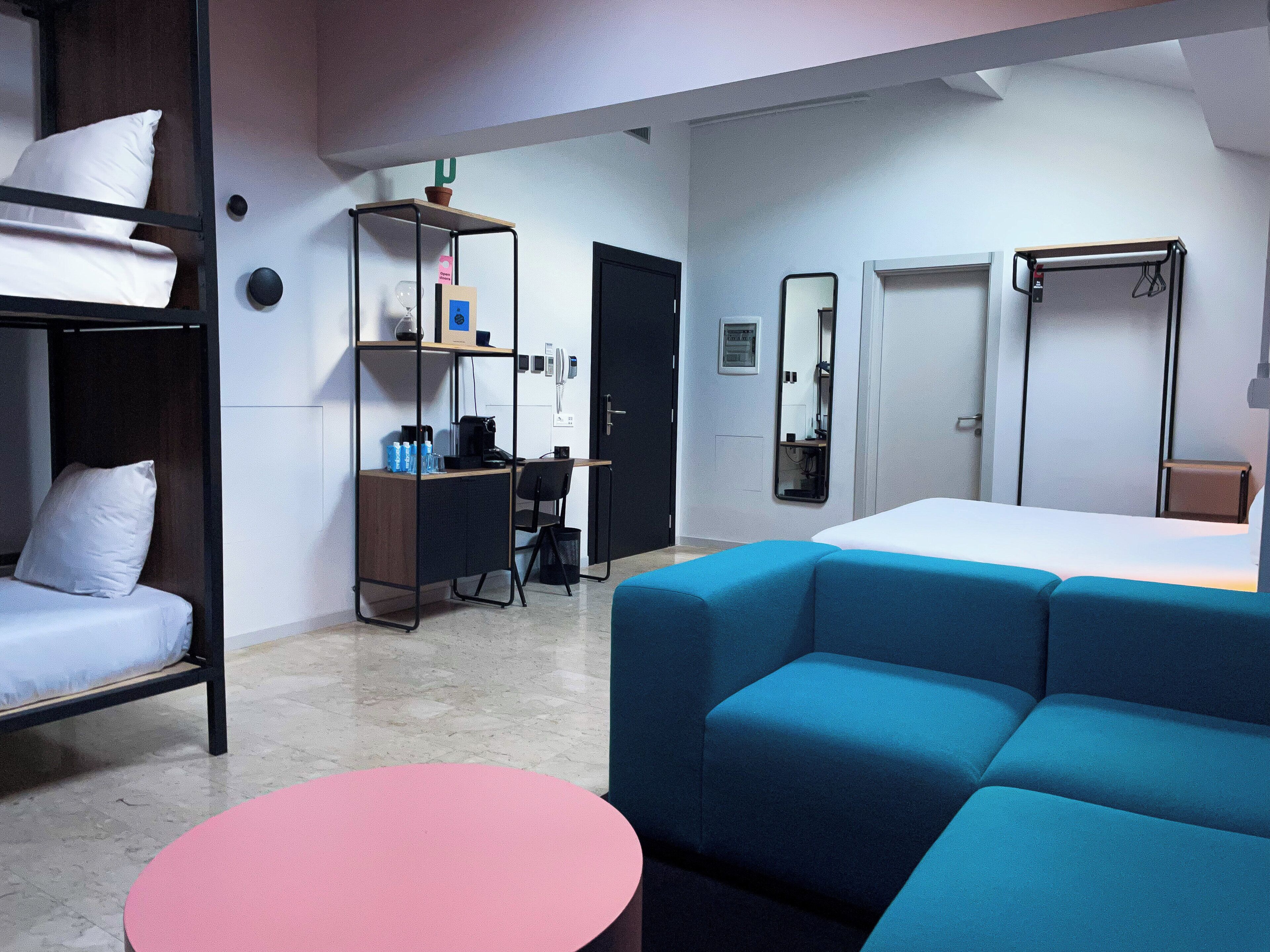 Ein Hotelzimmer mit Etagenbett, Schreibtisch mit Stuhl, einem kleinen runden Tisch, einer Couch, einem Spiegel und einem Kleiderschrank.