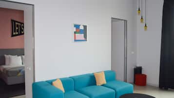 Quarto moderno com sofá azul, cama com roupa de cama branca, mesa pequena e quadro abstrato na parede.