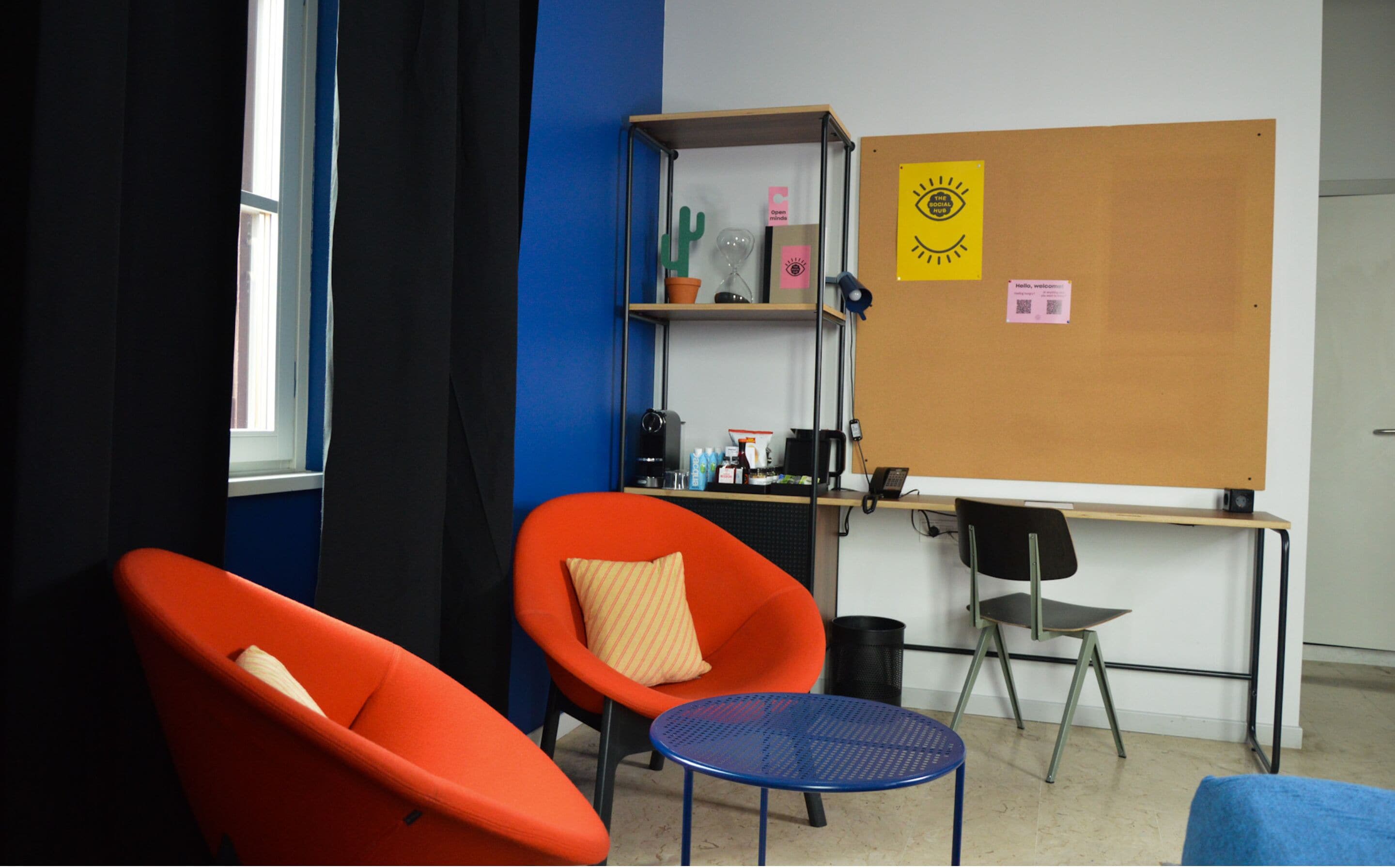 Ein Zimmer mit zwei orangefarbenen Stühlen, einem kleinen blauen Tisch, einem Schreibtisch mit einem Computer, einem Bücherregal und einer Pinwand mit Notizen.