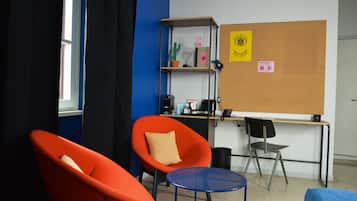 Ein Zimmer mit zwei orangefarbenen Stühlen, einem kleinen blauen Tisch, einem Schreibtisch mit einem Computer, einem Bücherregal und einer Pinwand mit Notizen.