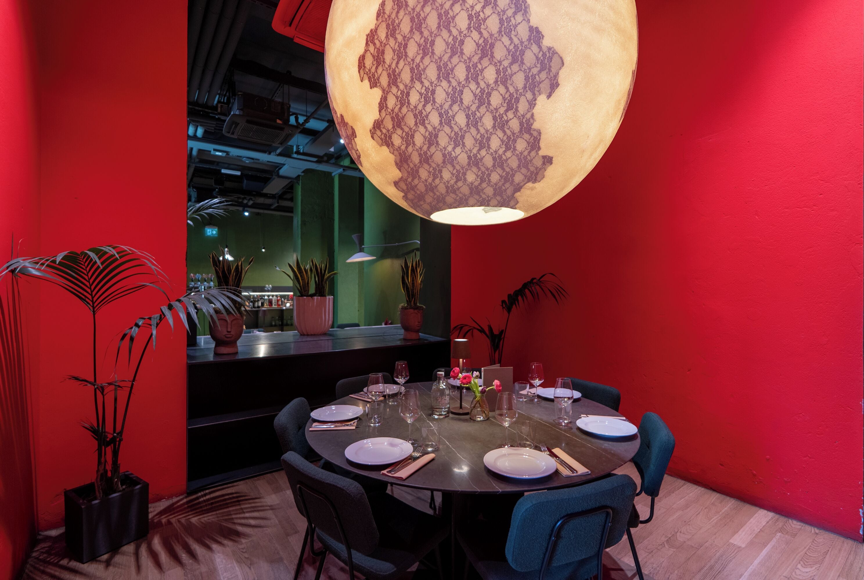 Zona pranzo con tavolo rotondo apparecchiato per quattro persone, circondato da sedie, con una parete rossa e un grande lampadario decorativo.