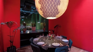 Zona pranzo con tavolo rotondo apparecchiato per quattro persone, circondato da sedie, con una parete rossa e un grande lampadario decorativo.