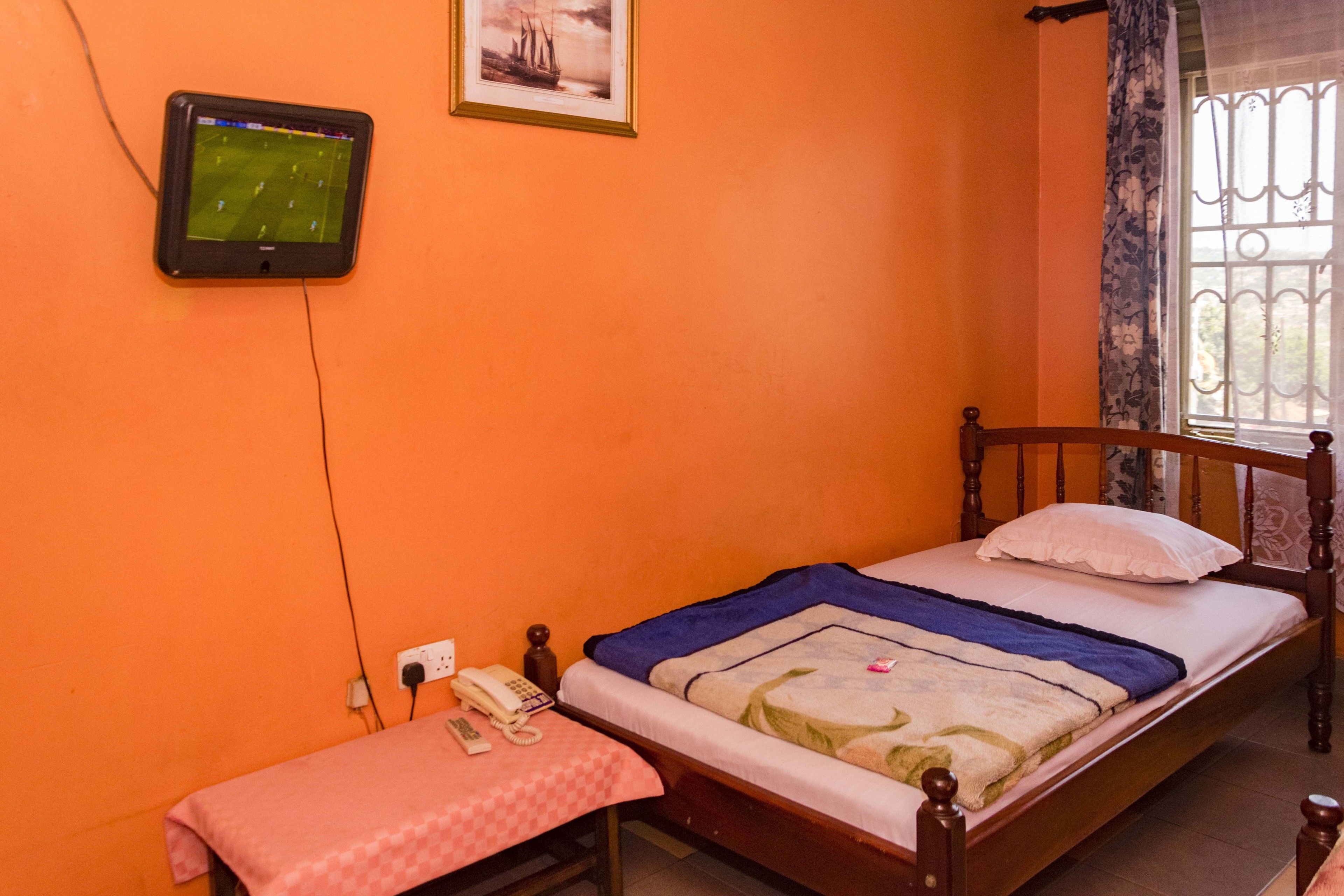 Een slaapkamer met een oranje wand, een houten bed, een klein nachtkastje, een aan de muur bevestigde televisie en een raam met gordijn.
