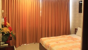 A hotel room with a bed, curtains, a desk with a vase of flowers, and a framed picture on the wall.