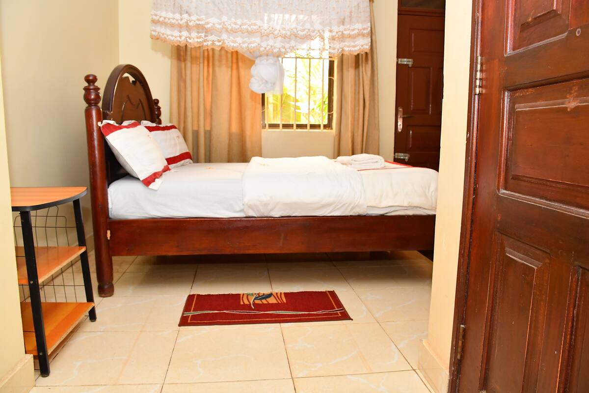 A wooden bed with white bedding, a red and yellow rug, and a wooden nightstand with a shelf.