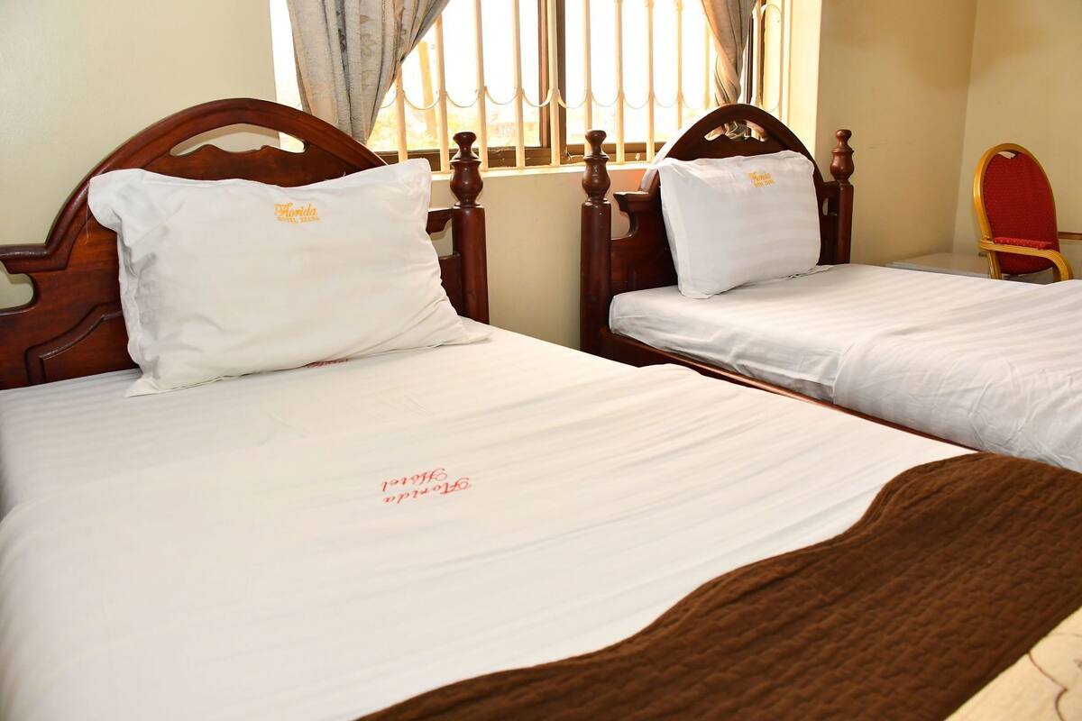 Two wooden beds with white linens and pillows, a red chair, and a window with vertical blinds.
