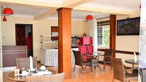A dining area with wicker chairs and tables, a kitchen area with red cabinets, and a TV mounted on the wall. - Florida Hotel (Kampala)