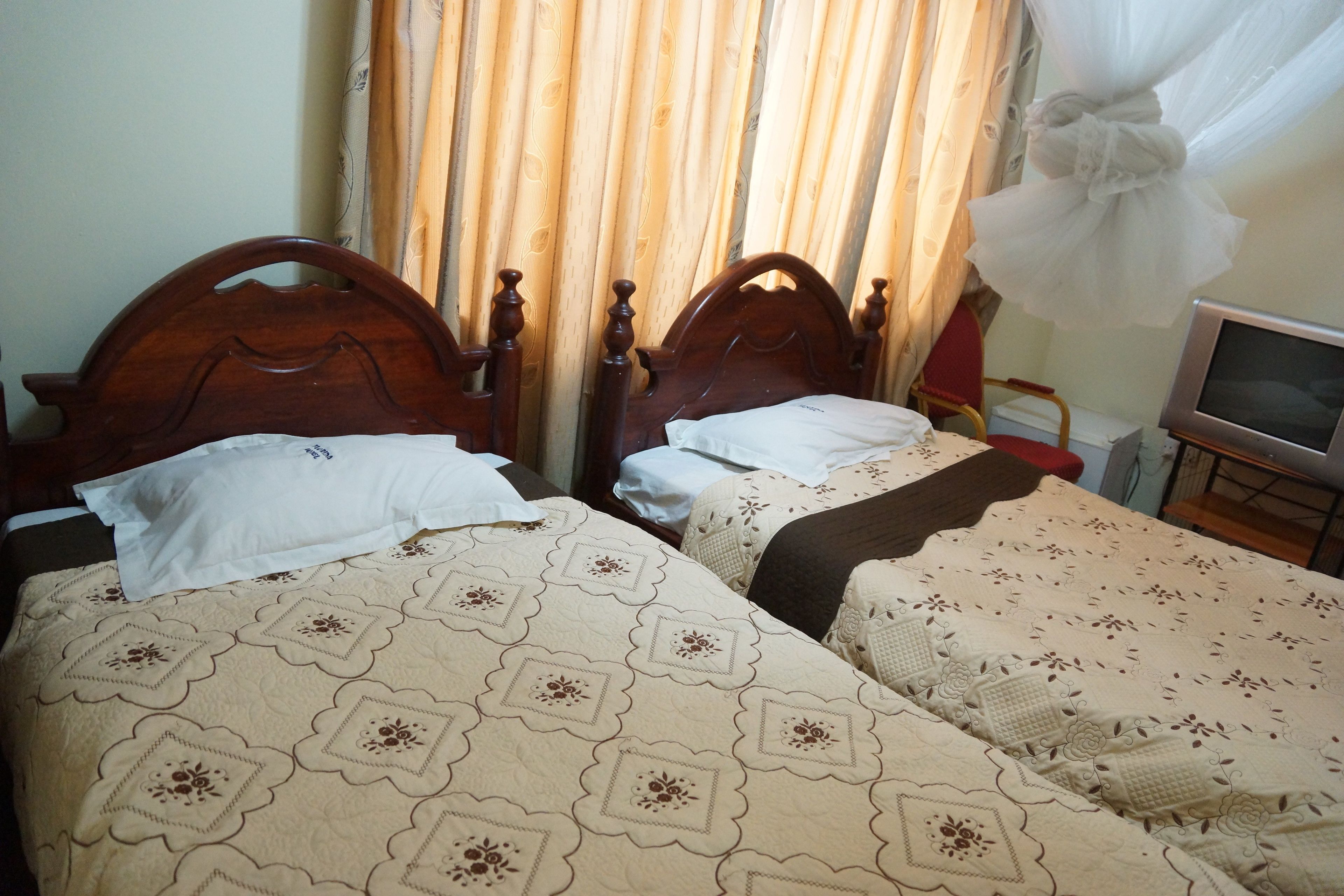 Duas camas de madeira com roupa de cama florida, uma televisão em um suporte e uma cortina com padrão de folhas.