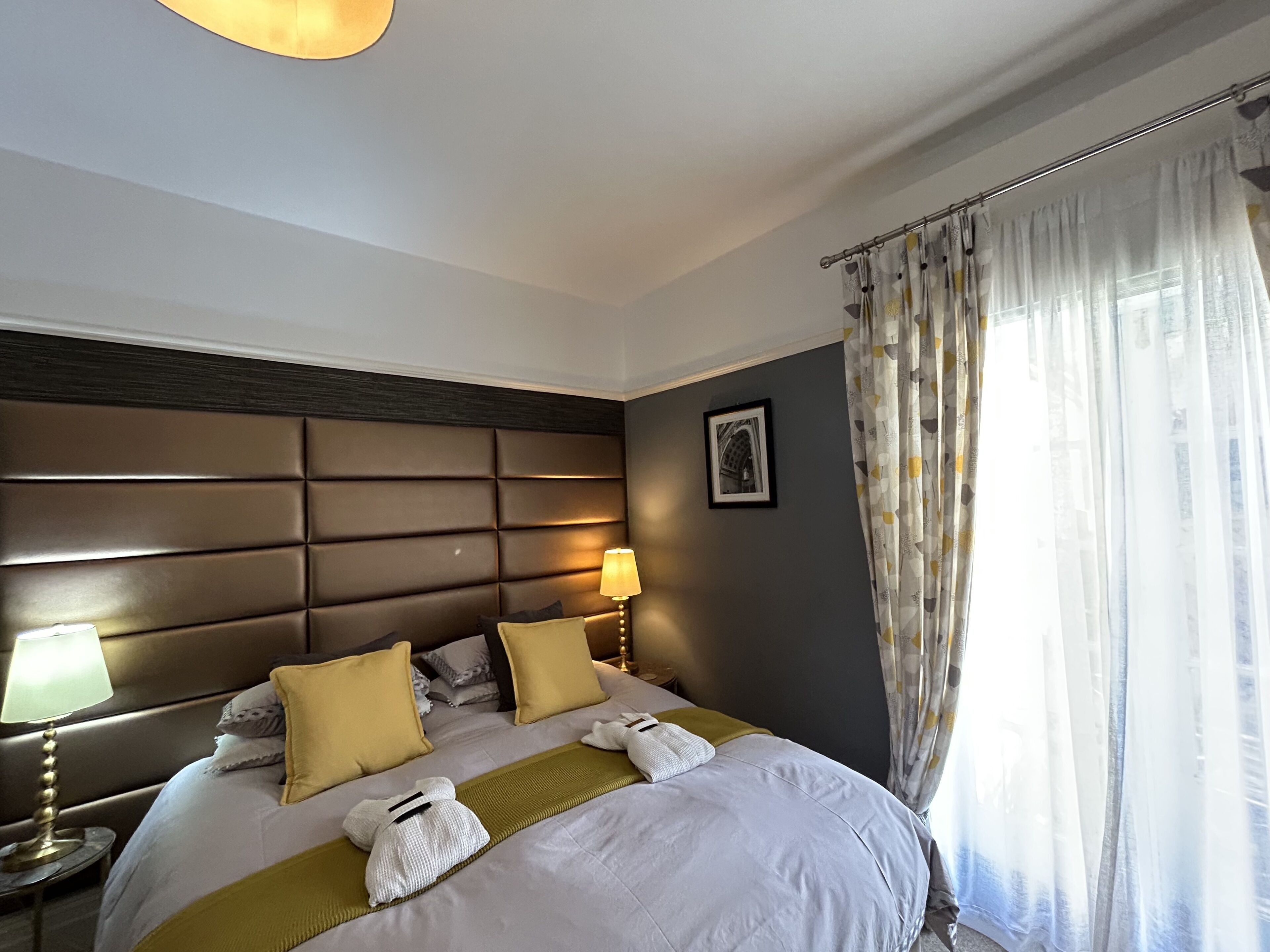 Ein Hotelzimmer mit einem großen Bett, einem Kopfteil, einem Nachttisch mit Lampe und einem Fenster mit Vorhängen.