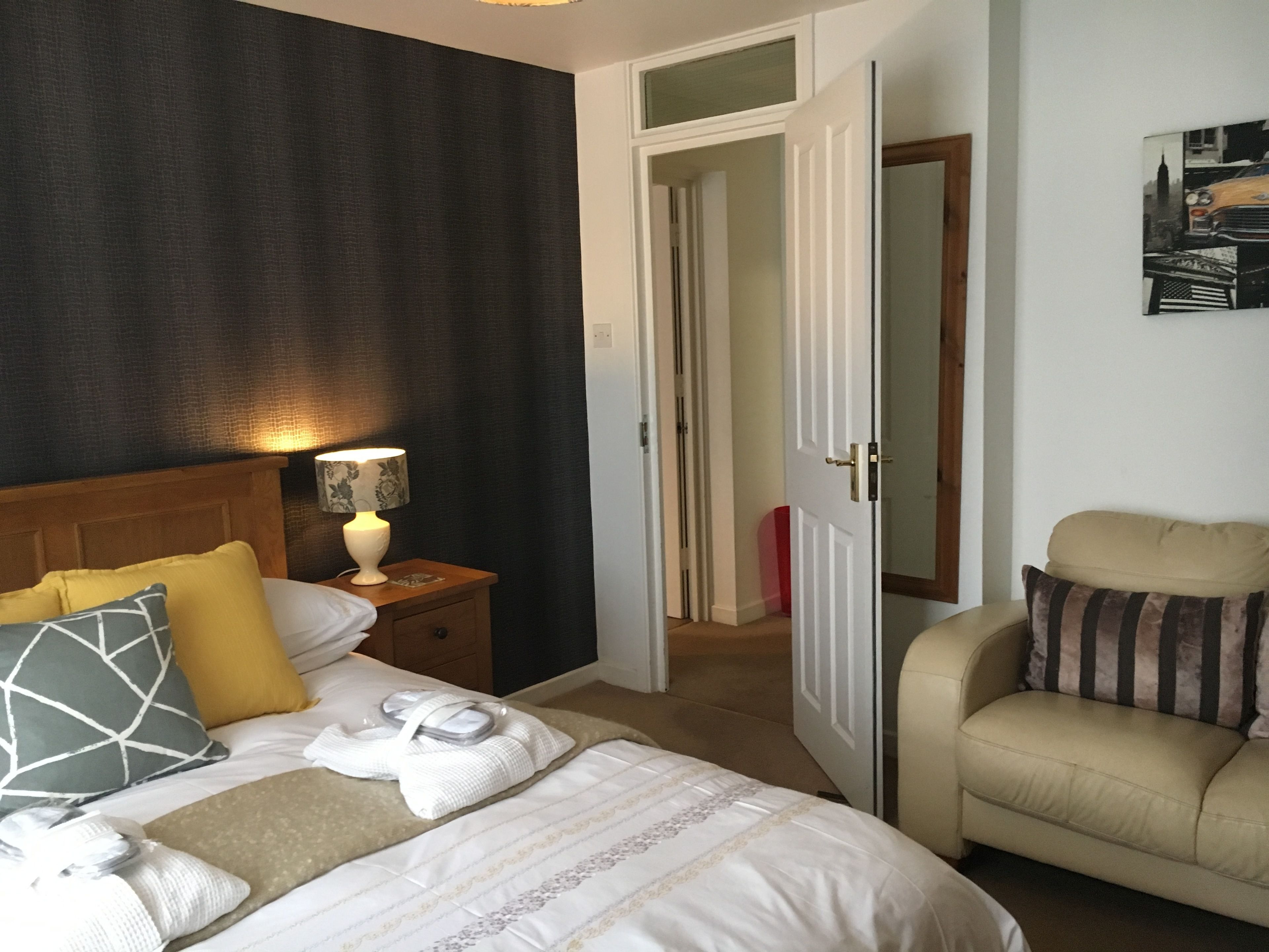 A hotel room with a bed, a nightstand with a lamp, a chair, and a door leading to another room.
