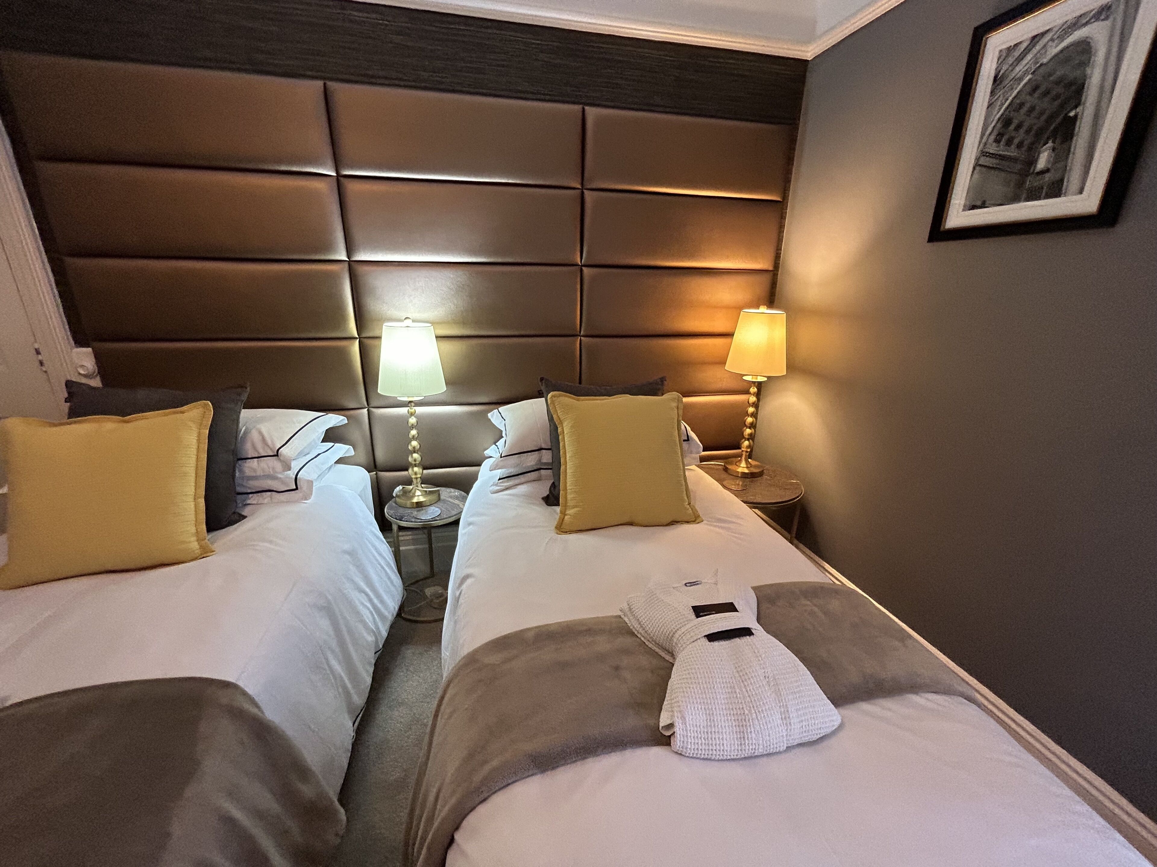 Une chambre d’hôtel avec deux lits, une tête de lit rembourrée, des lampes de chevet et un tableau encadré au mur.