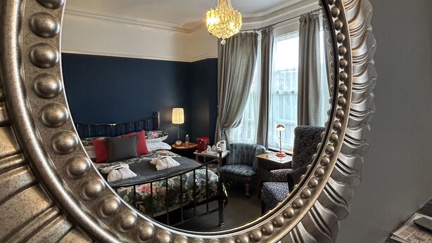 Een kamer met een grote, versierde spiegel, een kroonluchter, een bed met rode en grijze kussens, een stoel en een tafel met een lamp.