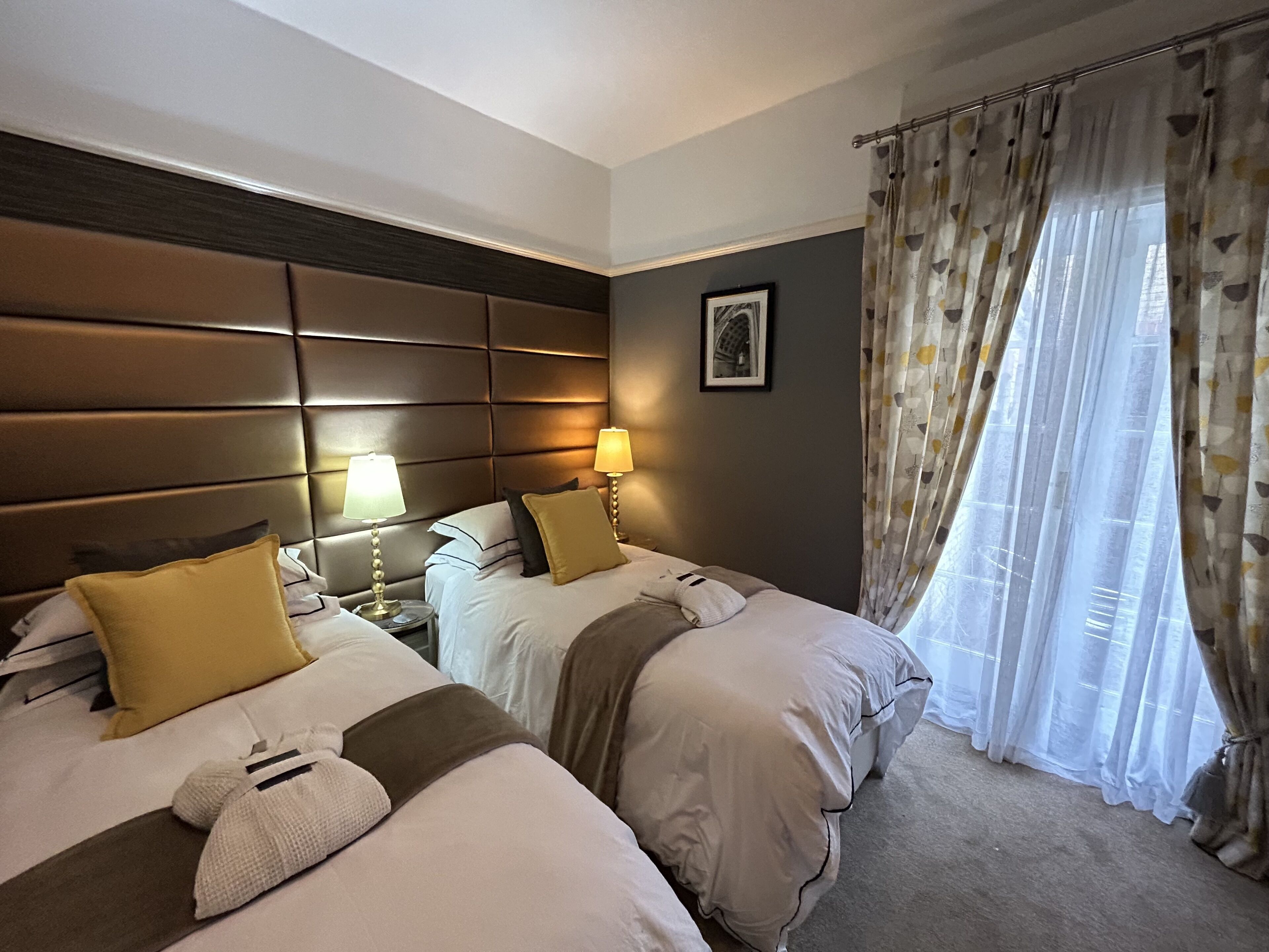 A hotel room with two beds, a headboard, bedside lamps, a framed picture, and a window with curtains.