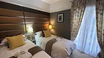 A hotel room with two beds, a headboard, bedside lamps, a framed picture, and a window with curtains.