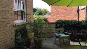 Een bakstenen gebouw met een raam, een bloempot, een houten stoel en een tafel onder een groene parasol.