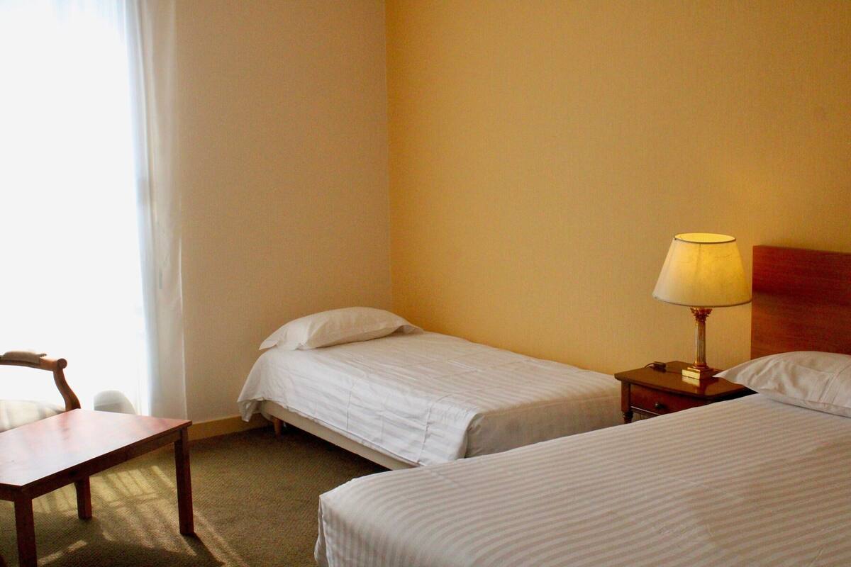 Habitación de hotel con dos camas individuales, una mesita de noche de madera con lámpara, una pequeña mesa y una silla.
