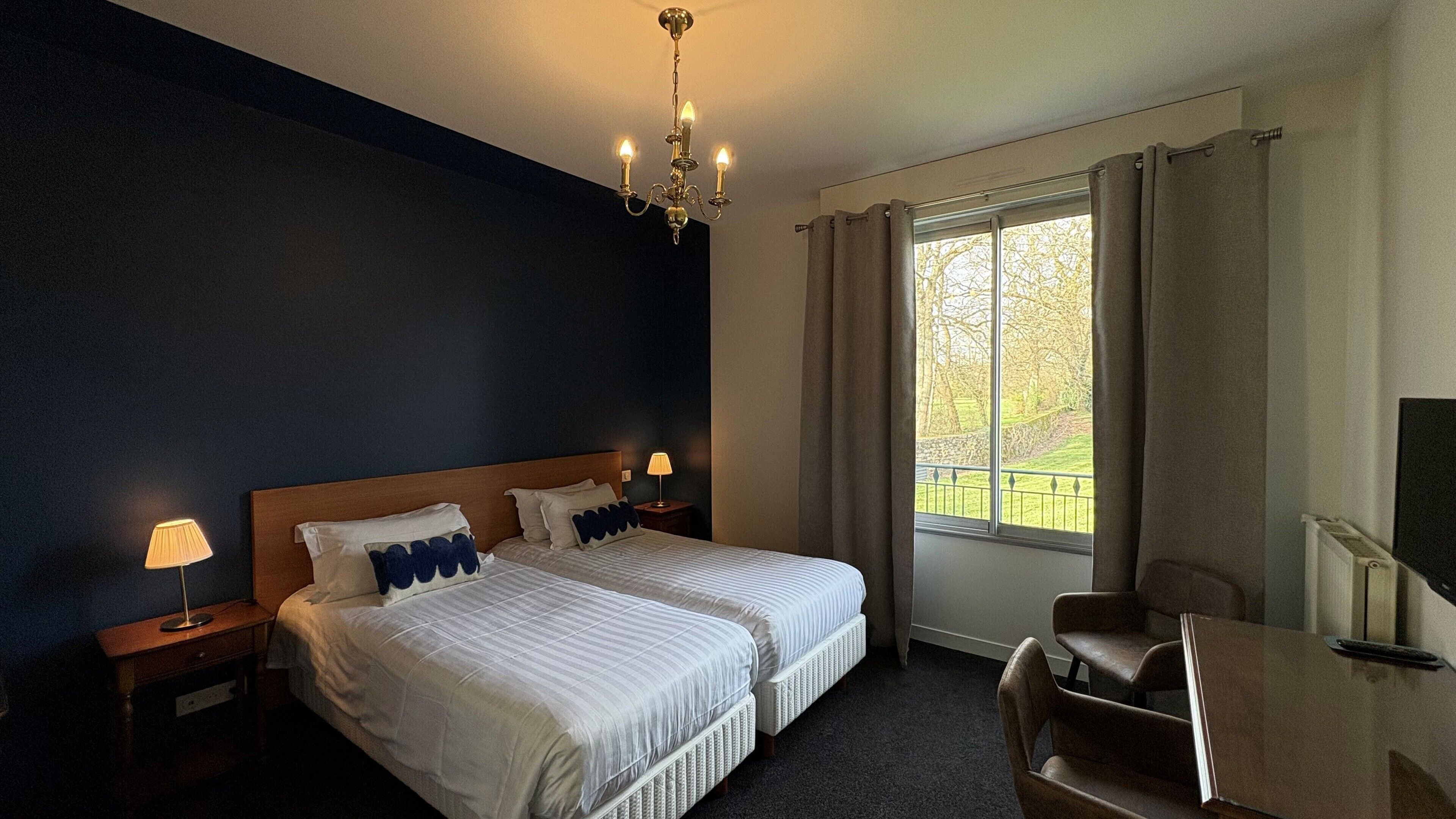 A hotel room with two beds, a chandelier, a desk with a chair, and a window with curtains.