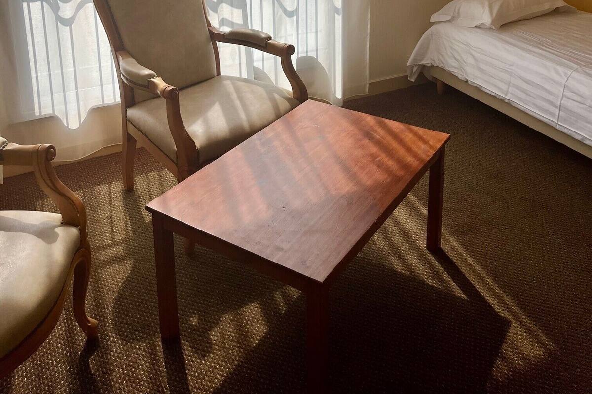 Una habitación con una silla de madera, una mesita pequeña de madera, una cama y cortinas traslúcidas.
