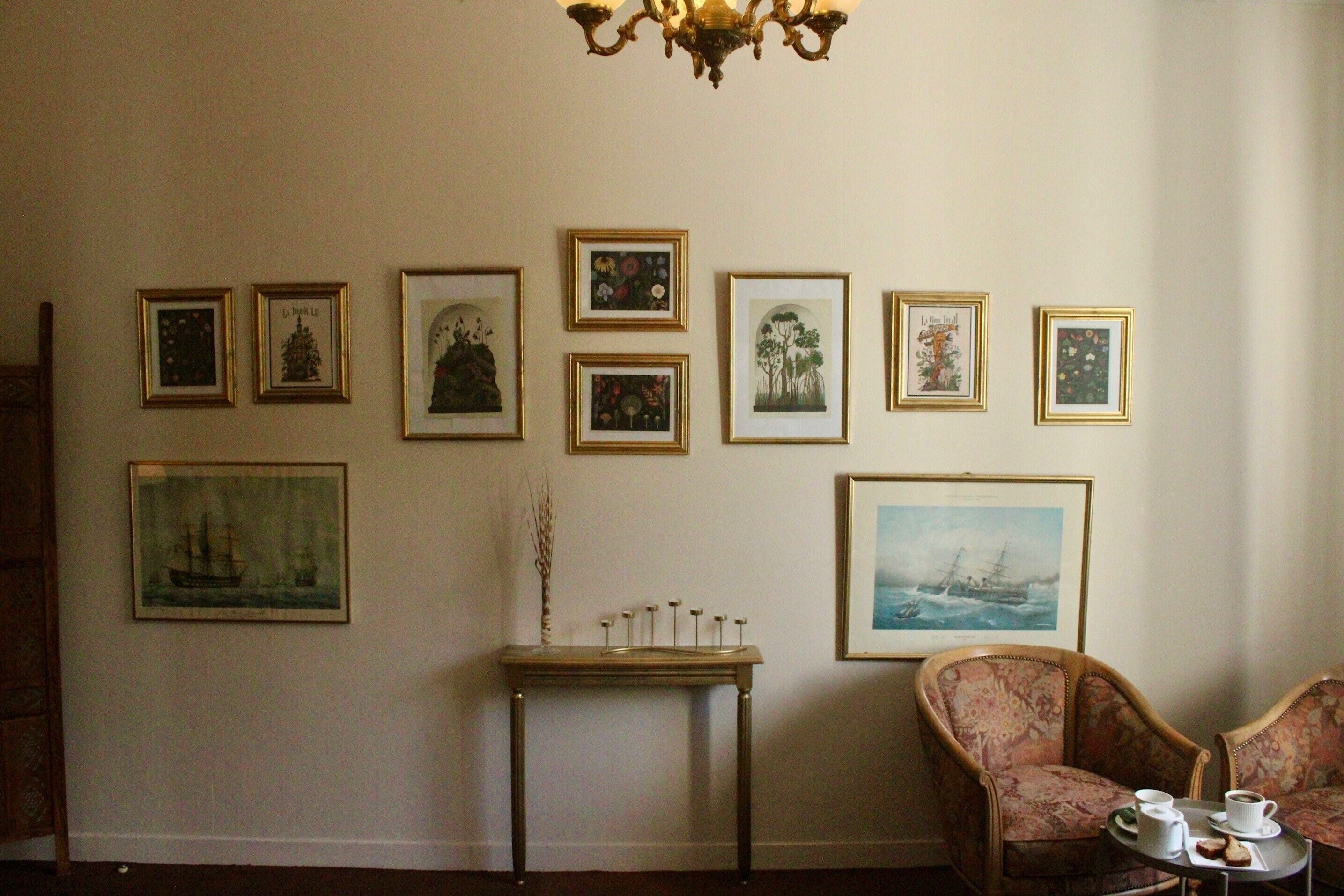 Ein Zimmer mit einer Bildergalerie an der Wand, einem kleinen Tisch mit einer Vase und Kerzen sowie zwei Polstersesseln mit Serviertablett.