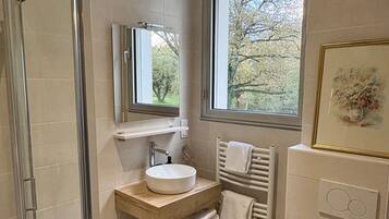 A modern bathroom with a glass shower enclosure, a wooden vanity, a white sink, a mirror, a window with a view of trees, and a toilet.