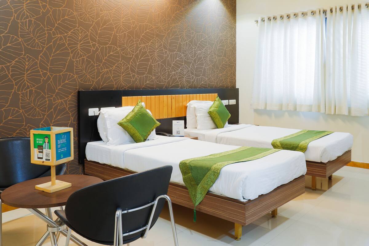 Une chambre d’hôtel avec deux lits, une tête de lit en bois, un bureau avec une chaise, une petite table et un mur orné d’un motif de feuilles.