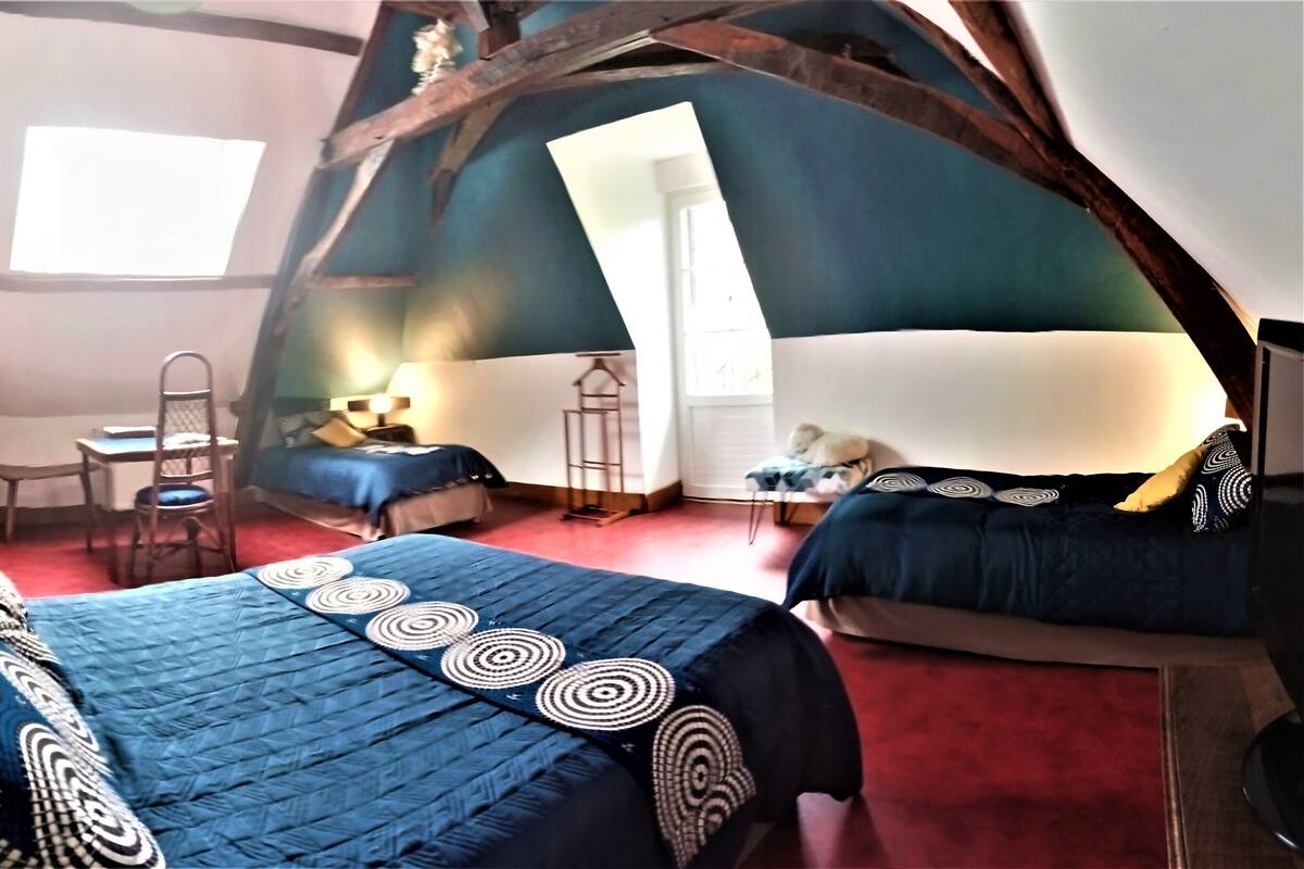 A cozy, rustic room with a large bed, a smaller bed, a desk, and a chair. There is a window providing natural light.