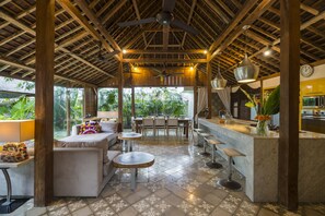 A spacious indoor area with a high ceiling, wooden beams, a dining table with chairs, a bar area, and a seating area with sofas and round tables. - Villa Batik Seminyak (Seminyak)
