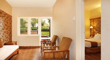 A hotel room with a bed, a wooden chair, a small table with a vase, and a window with a view of a garden.
